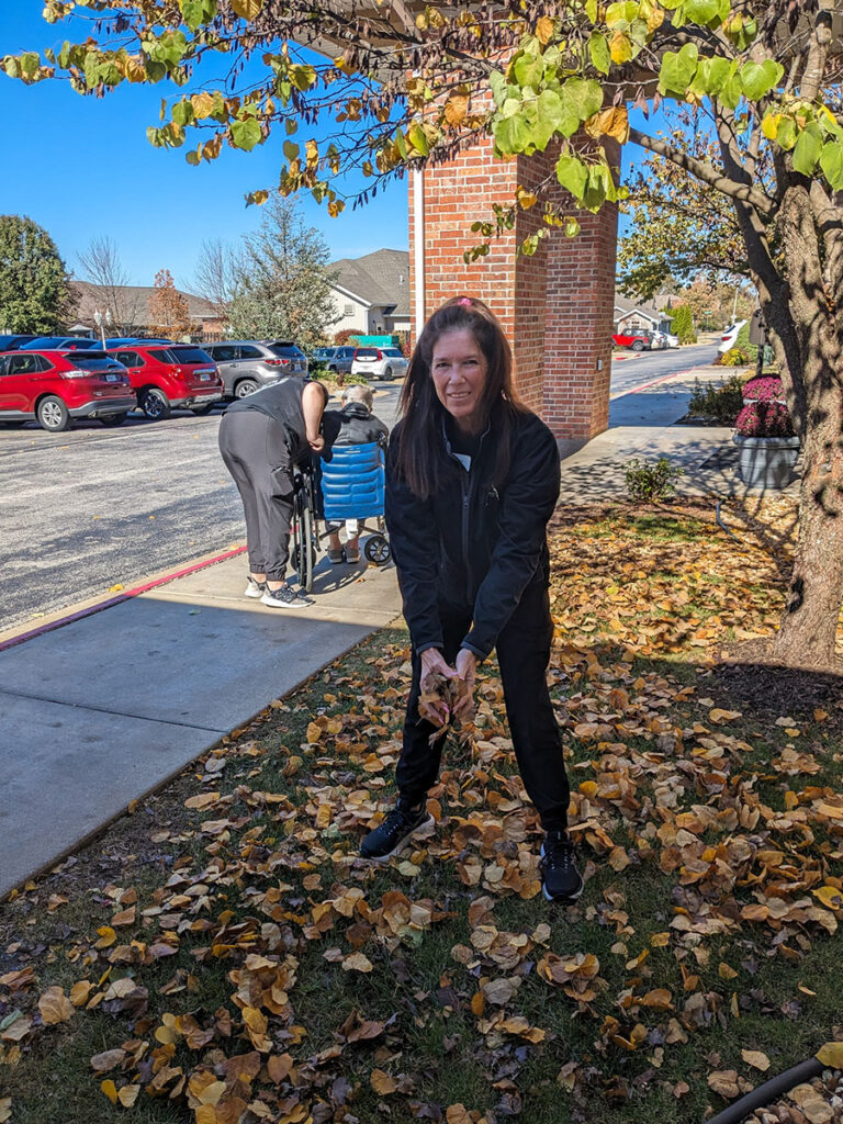 A woman in a black jacket holds a leaves while doing yard clean up, on a cool sunny day.