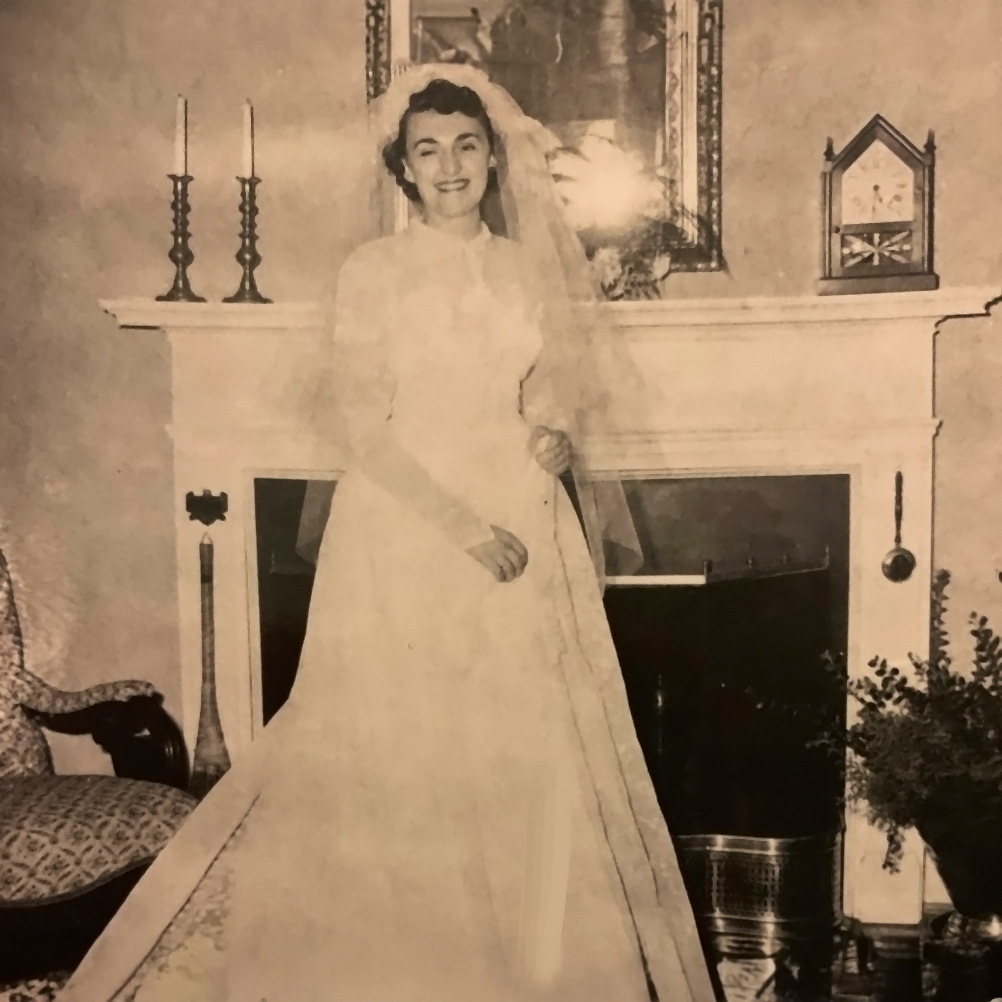 A vintage photo of Mary as a bride in a white wedding gown, standing inside by a fireplace, exudes timeless elegance and romance, preserving a cherished moment.