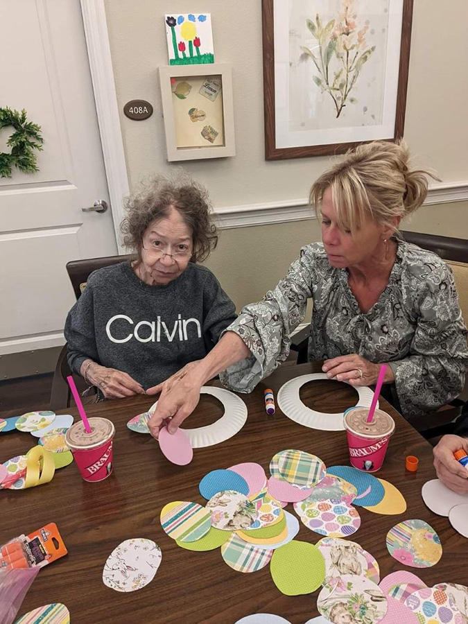 Two women, including Danielle, crafting paper at a table in memory care, making Easter wreaths with residents while volunteering.