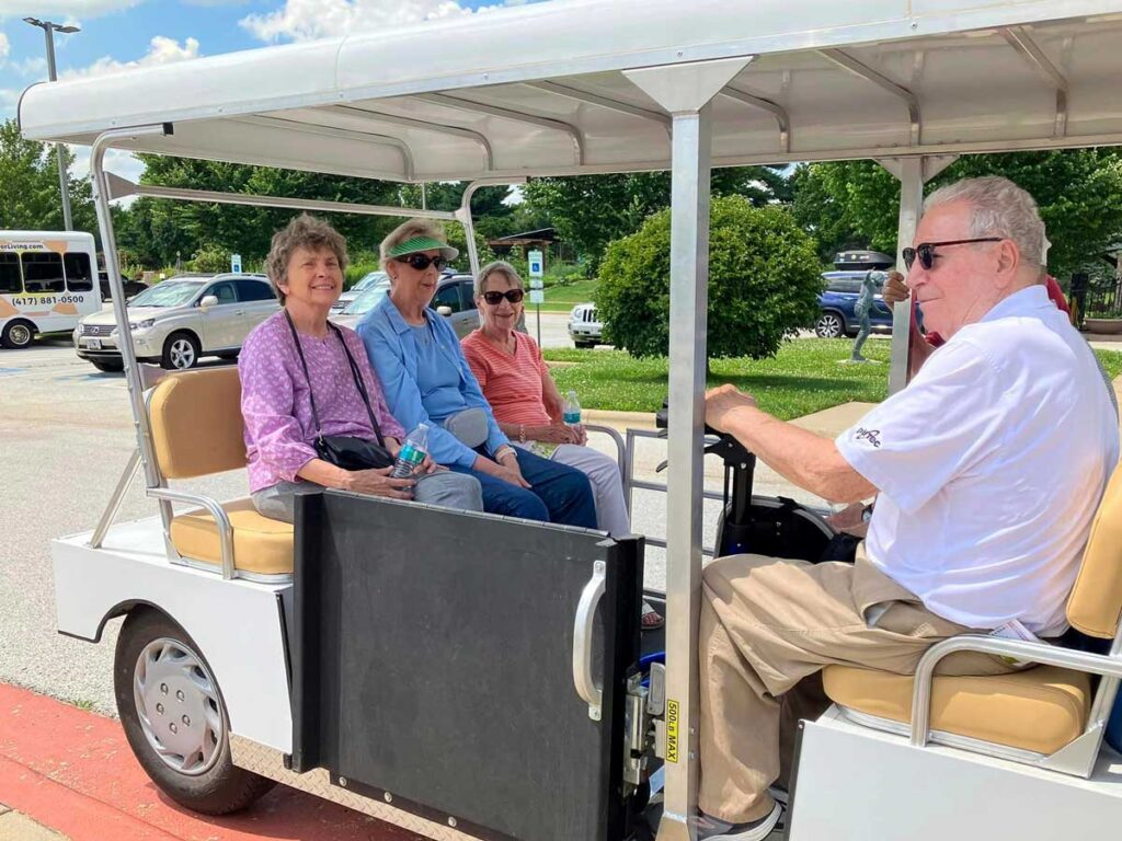 Group of senior residents sitting in a golf cart with a driver, enjoying an outing at a park on a sunny day.