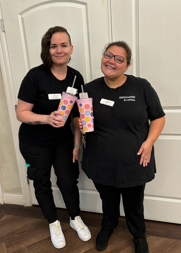 Two staff members at a senior living community smile while holding matching pink tumblers decorated with colorful smiley faces. They stand side by side, both wearing black uniforms, creating a cheerful and friendly atmosphere.