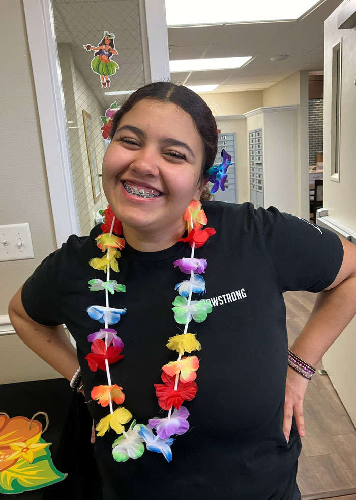 A smiling staff member at a senior living community wears a colorful flower lei and matching hair accessories, celebrating a tropical-themed event. Her cheerful expression and vibrant attire create a joyful and festive atmosphere.