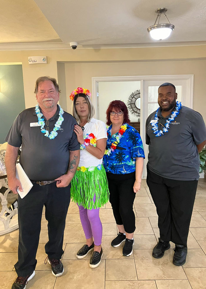 Four staff members at a senior living community pose together, dressed in tropical-themed attire, including leis, a grass skirt, and Hawaiian shirts. They are celebrating a themed event, creating a fun and festive atmosphere.