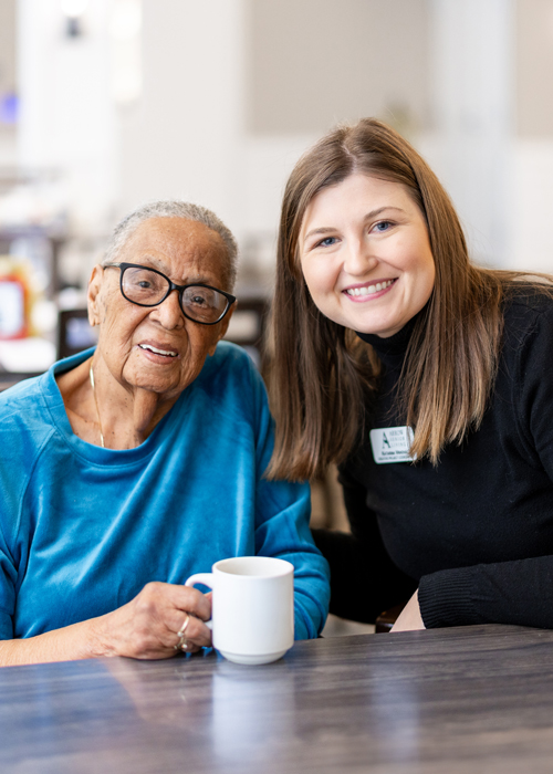 Senior resident in a blue sweater enjoys a cup of coffee while smiling alongside a friendly caregiver, emphasizing warmth, care, and companionship in the senior living community.