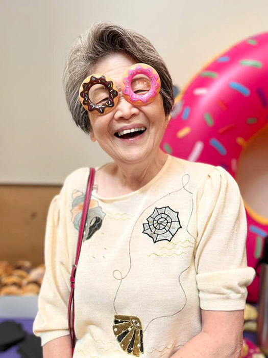 Senior resident joyfully wears playful donut-shaped glasses, standing in front of a large inflatable donut with sprinkles, highlighting a fun and lighthearted atmosphere.