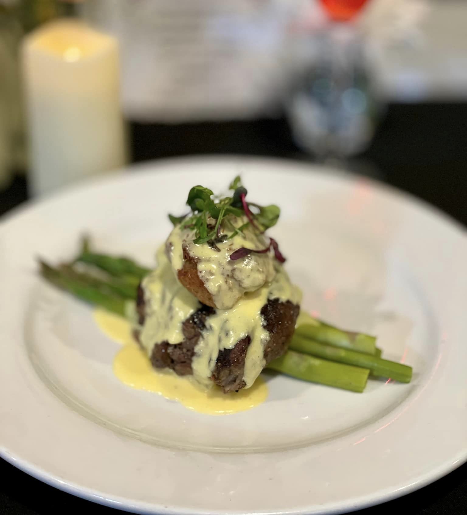 Plated gourmet steak with asparagus, topped with creamy hollandaise sauce and garnished with microgreens, set against a blurred background of a candle, flowers, and drink.