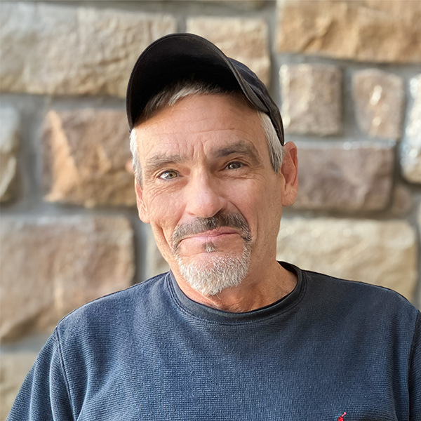 Darin Maples, Plant Operations Director at The Fremont Senior Living, smiles warmly in a casual cap and sweater against a stone wall, reflecting his dedication to the community.