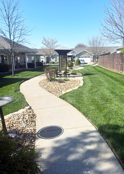 Sunny spring day at The Fremont Senior Living with a curved walking path, green lawn, and shaded seating area between cottage-style homes.