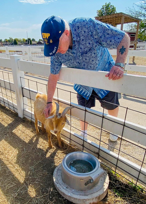 Senior resident wearing a U.S. Navy cap gently pets a goat through a fence during a sunny outing to Rutledge-Wilson Farm Park, enjoying animal interaction and nature.