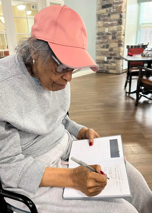 Senior resident wearing a pink hat and glasses writes in a gratitude journal, thoughtfully reflecting during a quiet moment in a cozy community space.
