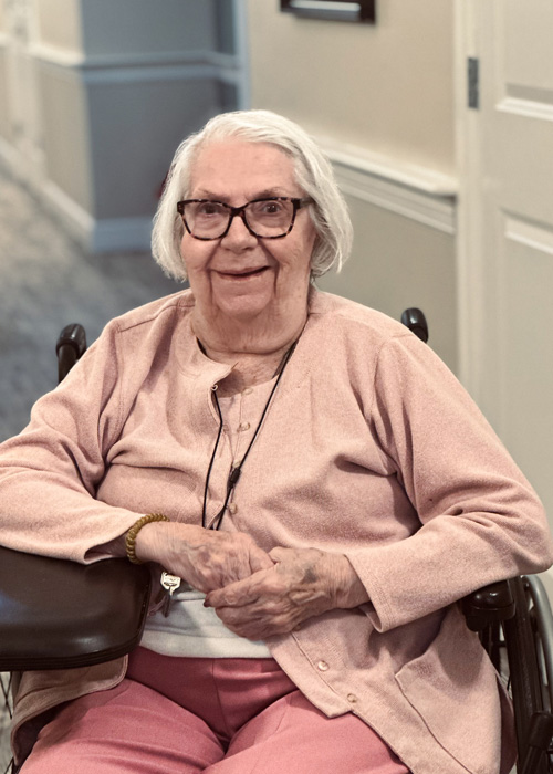 Senior resident in a wheelchair smiles warmly in a hallway, wearing a light pink sweater and enjoying a peaceful moment in the senior living community.