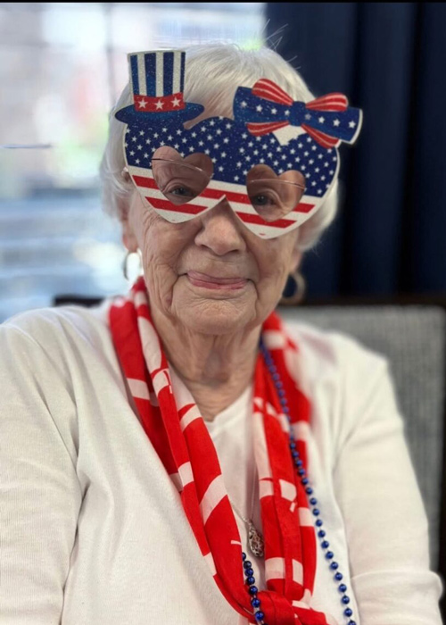 A senior woman wearing festive heart-shaped American flag glasses, a red and white scarf, and blue beads smiles during a Fourth of July celebration.