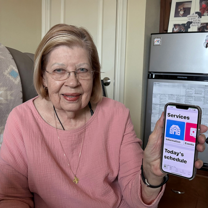 Smiling senior resident wearing a pink top, holding a smartphone that displays an app screen with icons labeled "Information," "Events," and "Today's schedule."