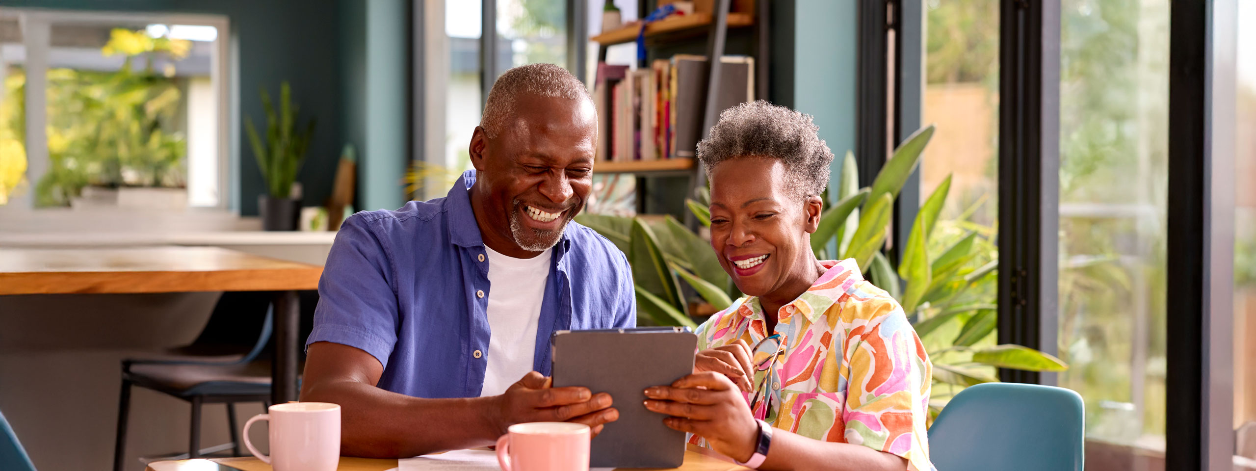 Happy senior couple sitting at a table with coffee mugs, smiling while using a digital tablet together in a bright, modern room filled with plants.