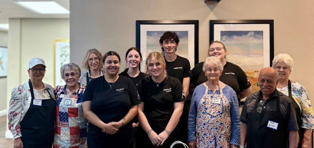 Senior living residents and team members stand together smiling in a group photo, some wearing aprons, showcasing community, teamwork, and connection during a dining event.