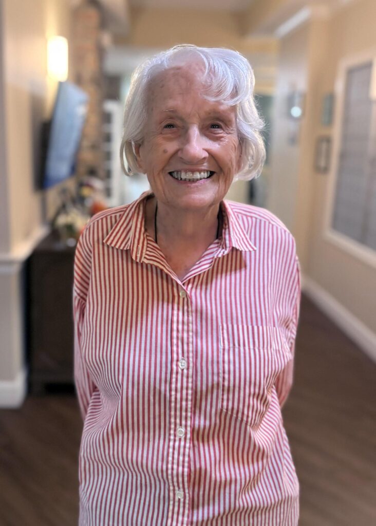 A senior woman in a button down long sleeved shirt smiles, hands clasped behind her back, at The Fremont Senior Living.