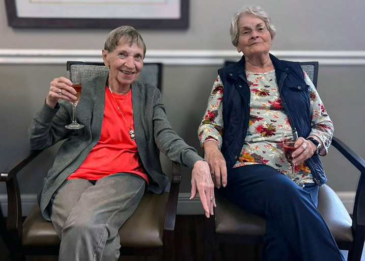 Two senior women enjoy a relaxed moment, seated side by side with drinks in hand.