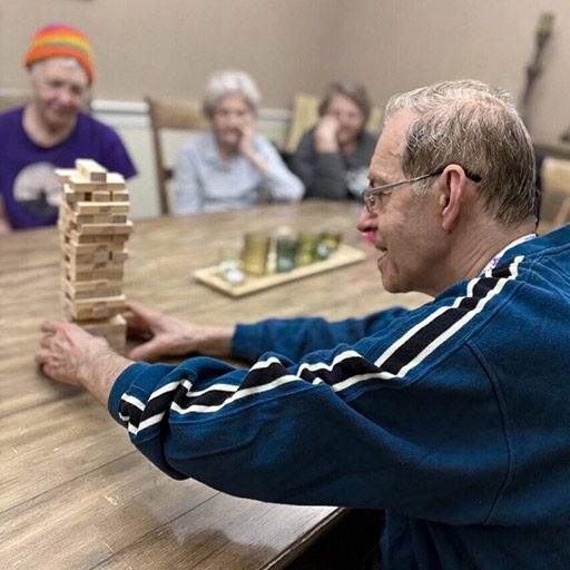 A senior man places a block on a Jenga tower while fellow residents look on.