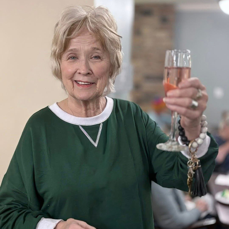 A senior woman in a green sweater smiles while raising a delicate drink in toast.