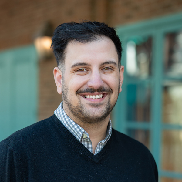 Anthony Argenta, Wellness Director at The Fremont Senior Living, smiling in a professional outdoor headshot with dark hair, a trimmed beard, and softly blurred brick and teal window details in the background.