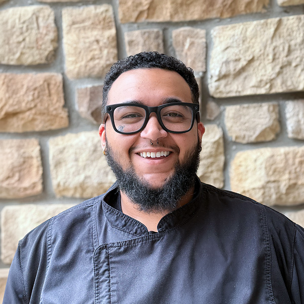 Devon Gardner, Culinary Director at The Fremont Senior Living, smiling in a professional headshot with black glasses, a short curly haircut, a beard, and a light stone wall background.