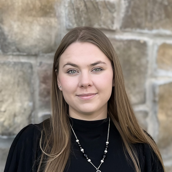 Sierra Jewell, Senior Living Coordinator at The Fremont Senior Living, looking at the camera in a professional headshot with long straight light brown hair, a black top, a pendant necklace, and a stone wall background.