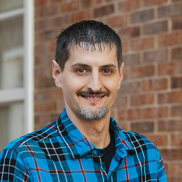 Wyatt Koory, Plant Operations Director at The Fremont Senior Living, smiling in a professional outdoor headshot with short dark hair, a trimmed goatee, a blue plaid button-up shirt, and a softly blurred brick background.