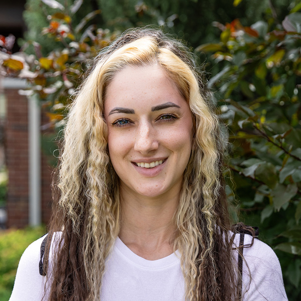 Morgan Carter, Memory Care Director at The Fremont Senior Living, smiling in a professional outdoor headshot with long crimped blonde and brown hair, a white top, and a softly blurred green foliage background.
