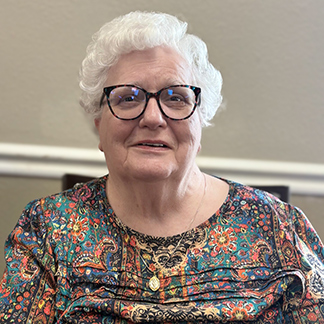 A senior woman wearing a colorfully patterned blouse and glasses smiles at The Fremont Senior Living.