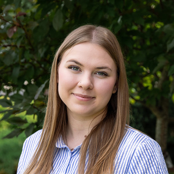 Sierra Jewell, Senior Living Coordinator at The Fremont Senior Living, looking at the camera in a professional outdoor headshot with long straight light brown hair, a blue striped button-up shirt, and a softly blurred green foliage background.