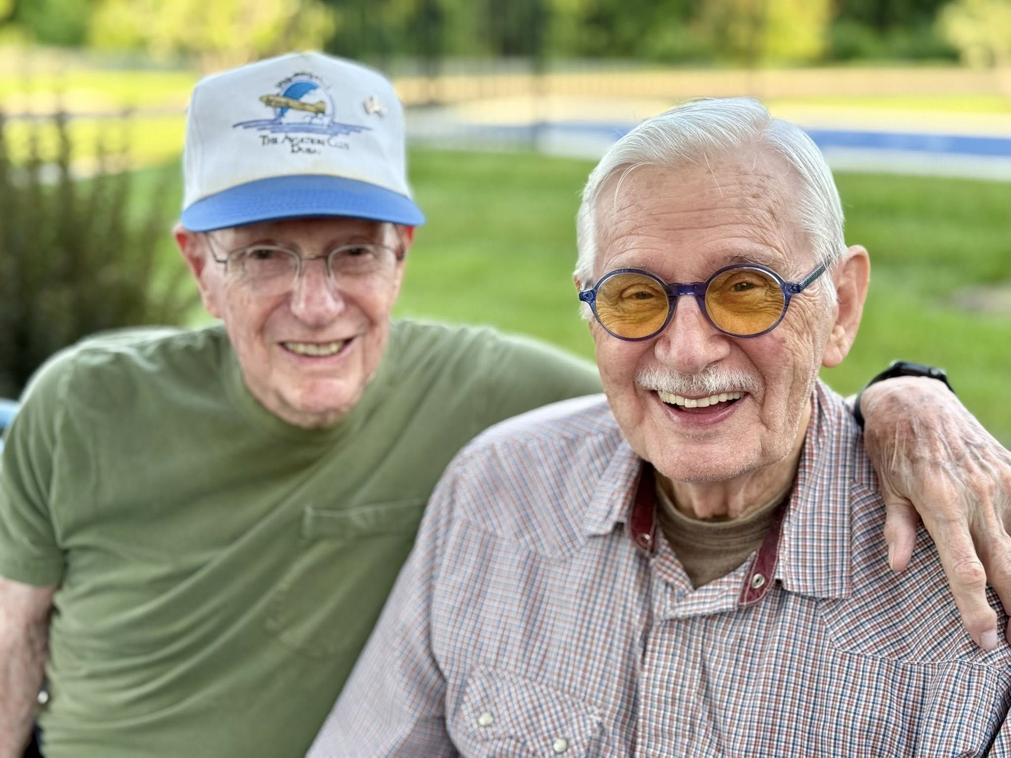 Two older men smiling together outdoors, with one resting his arm around the other in a sunny green setting.