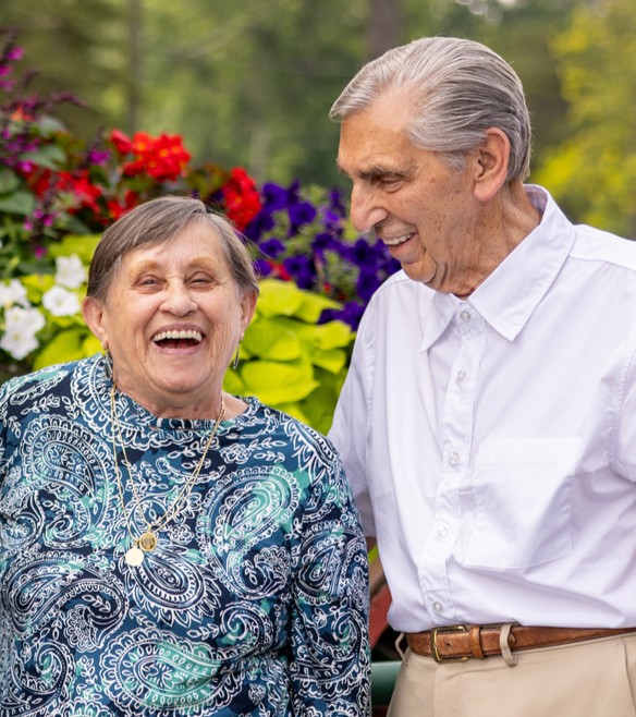 Older woman laughing beside an older man as they enjoy time together in a colorful outdoor garden.