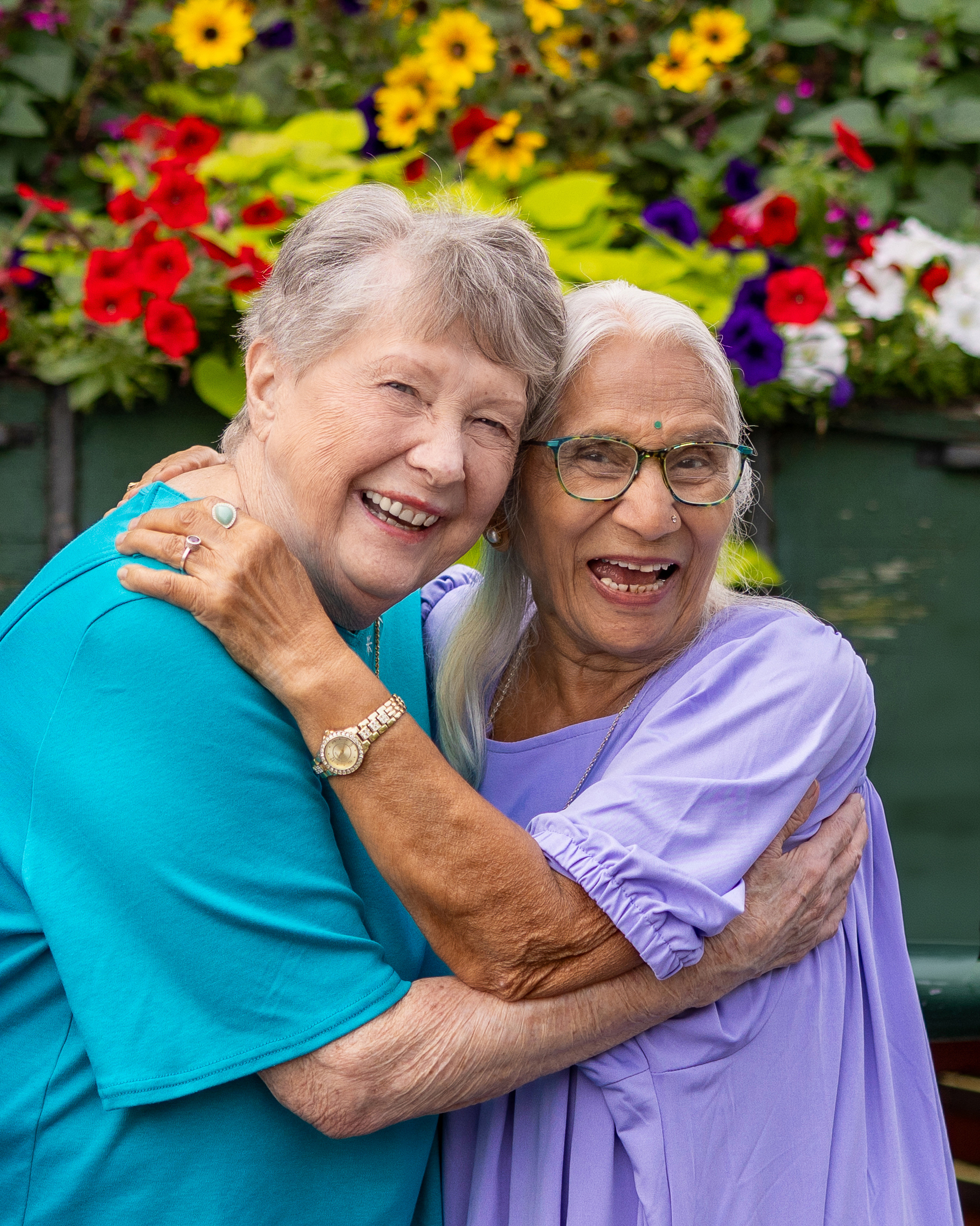 Two older women smiling and embracing in front of a colorful garden filled with bright flowers.